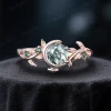 A side profile view of a 1 carat round cut natural moss agate ring set in 14k rose gold. The vintage design features a moon and star motif with small green emerald accents surrounding the leaf-green agate center stone.
