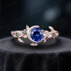 A side profile of a 1 carat round cut natural blue sapphire ring set in 14k rose gold. The vintage moon and star design features vibrant green emerald accents surrounding the deep blue sapphire center stone, with delicate leaf-inspired detailing along the band.