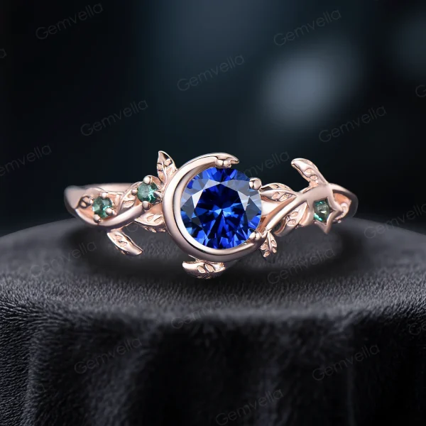 A side profile of a 1 carat round cut natural blue sapphire ring set in 14k rose gold. The vintage moon and star design features vibrant green emerald accents surrounding the deep blue sapphire center stone, with delicate leaf-inspired detailing along the band.