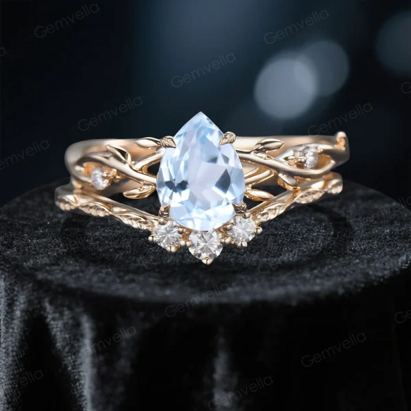 Close-up front view of a 1.25 carat pear cut aquamarine bridal ring set featuring a yellow gold band with detailed moissanite leaf accents, highlighting the center stone's icy blue color and the side stones' brilliance.