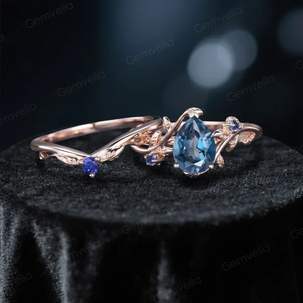 Flat lay display of the handmade bridal set, showing the 1.25ct pear-cut London blue topaz engagement ring and matching blue sapphire wedding band with intricate leaf-branch patterns in rose gold.