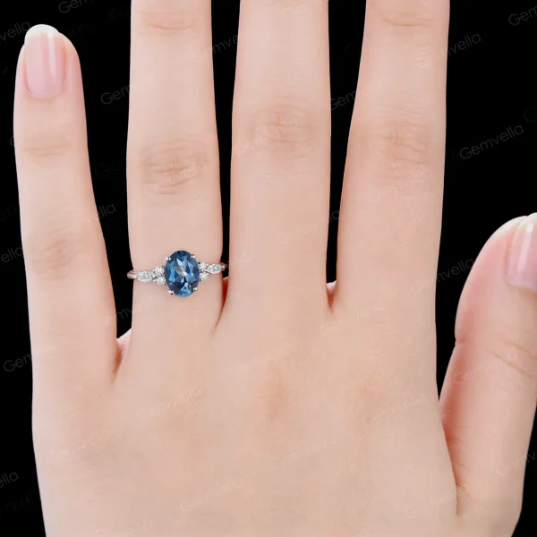 A woman's hand wearing the unique 1.5ct oval London Blue Topaz and marquise moissanite engagement ring. This handmade bridal jewelry makes a beautiful promise ring and anniversary gift for her, showing its elegant scale and sparkle on the finger.