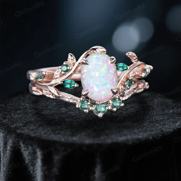 Angled view of a nature-inspired bridal ring set, highlighting the 1.5ct oval white opal center, 10K rose gold leaf vine branch design, emerald and moss agate accents on the engagement ring, and the matching emerald wedding band stacked below
