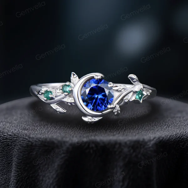 A delicate 925 sterling silver ring featuring a 1ct round blue sapphire stone. The design highlights a vintage moon and star theme with leaf green emerald accents, and subtle leaf engravings on the band for a cohesive nature-inspired aesthetic.
