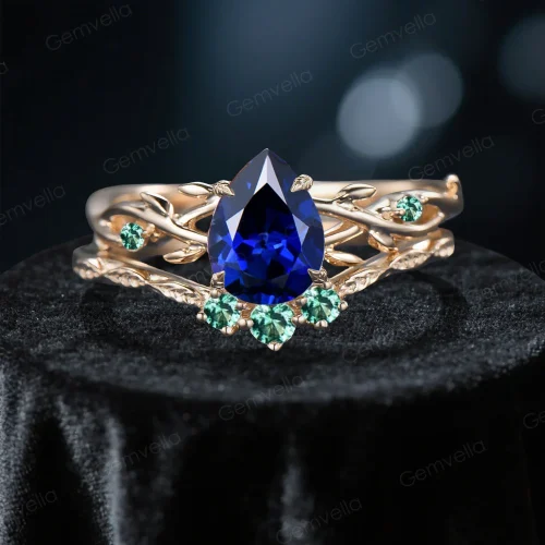 Close-up front view of a 1.25 carat pear cut blue sapphire engagement ring set featuring a yellow gold band with detailed green emerald leaf accents and organic twig textures surrounding the vivid blue center stone.