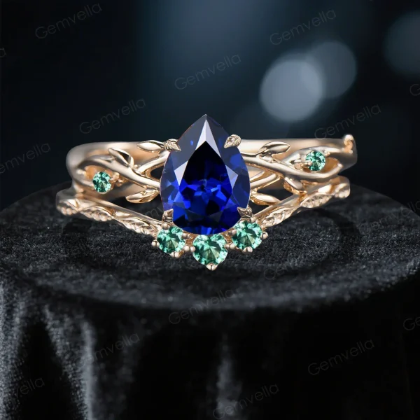 Close-up front view of a 1.25 carat pear cut blue sapphire engagement ring set featuring a yellow gold band with detailed green emerald leaf accents and organic twig textures surrounding the vivid blue center stone.