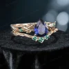 Side profile shot of the 1.25ct pear shaped blue sapphire ring on a yellow gold band, highlighting the prong setting height, the curvature of the matching wedding band, and the organic vine texture with emerald leaves.