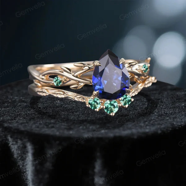 Side profile shot of the 1.25ct pear shaped blue sapphire ring on a yellow gold band, highlighting the prong setting height, the curvature of the matching wedding band, and the organic vine texture with emerald leaves.