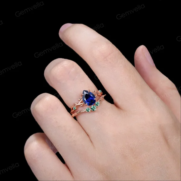 Lifestyle shot of a woman's hand wearing the 1.25ct pear shaped blue sapphire wedding set, showcasing how the nature inspired yellow gold twig ring with emerald leaves looks when worn and stacked on the finger.