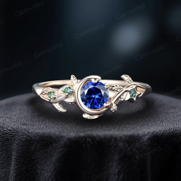 Close-up of a vintage style blue sapphire ring crafted in solid 14k yellow gold. The round cut blue sapphire is accented by genuine green emeralds in a celestial moon and star setting, with leaf motifs etched into the band for a nature-inspired look.