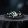 A striking side view of a 1ct round cut natural moss agate ring set in black gold. The vintage moon and star design features contrasting green emerald accents, creating a unique and modern nature-inspired look for women.