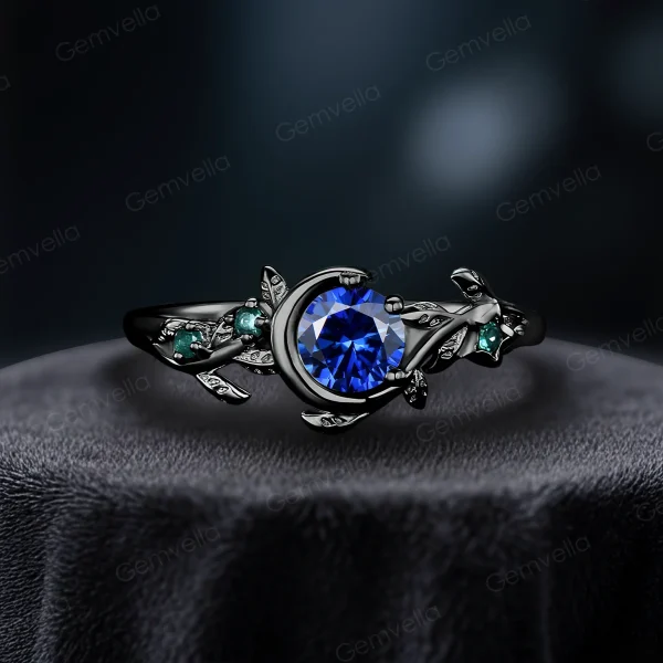A striking side view of a 1ct round cut natural blue sapphire ring set in 14k black gold. The vintage moon and star design features contrasting green emerald accents, with leaf-inspired detailing along the band to enhance the nature-themed look.